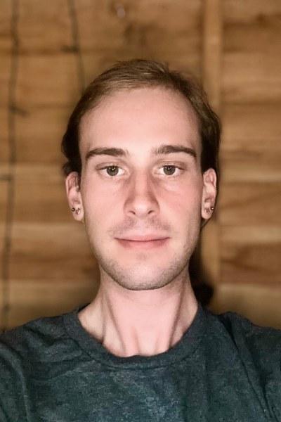 Bookkeeper Luke Wood wearing a grey top and hair tied back against a wooden background.