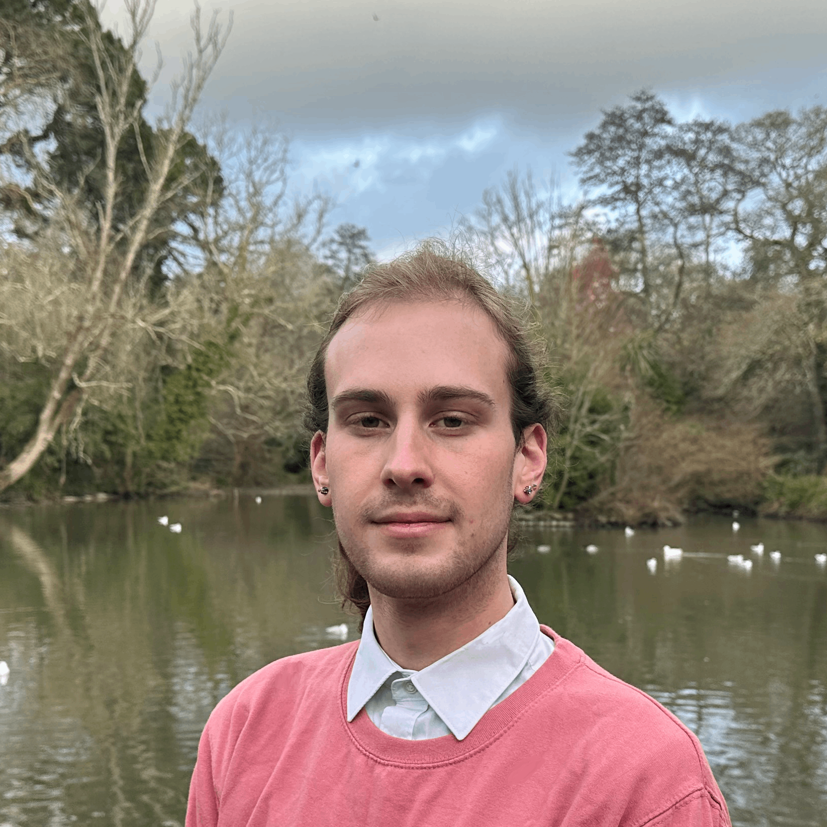 Photo of Luke Wood, owner of Luke Wood Bookkeeping, wearing a Pink jumper and white collared shirt, standing in front of a pond and trees in a park in Truro